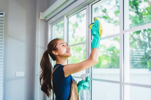 Young Asian housekeeper cleans a large window with a yellow cloth, smiling brightly in natural sunlight.